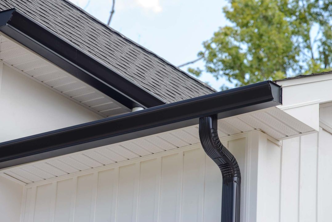 Black gutter and downspout on a white house roofline against a blue sky