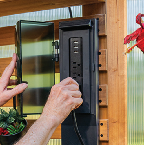 Hands installing a black outdoor electrical outlet box on a wooden wall panel.