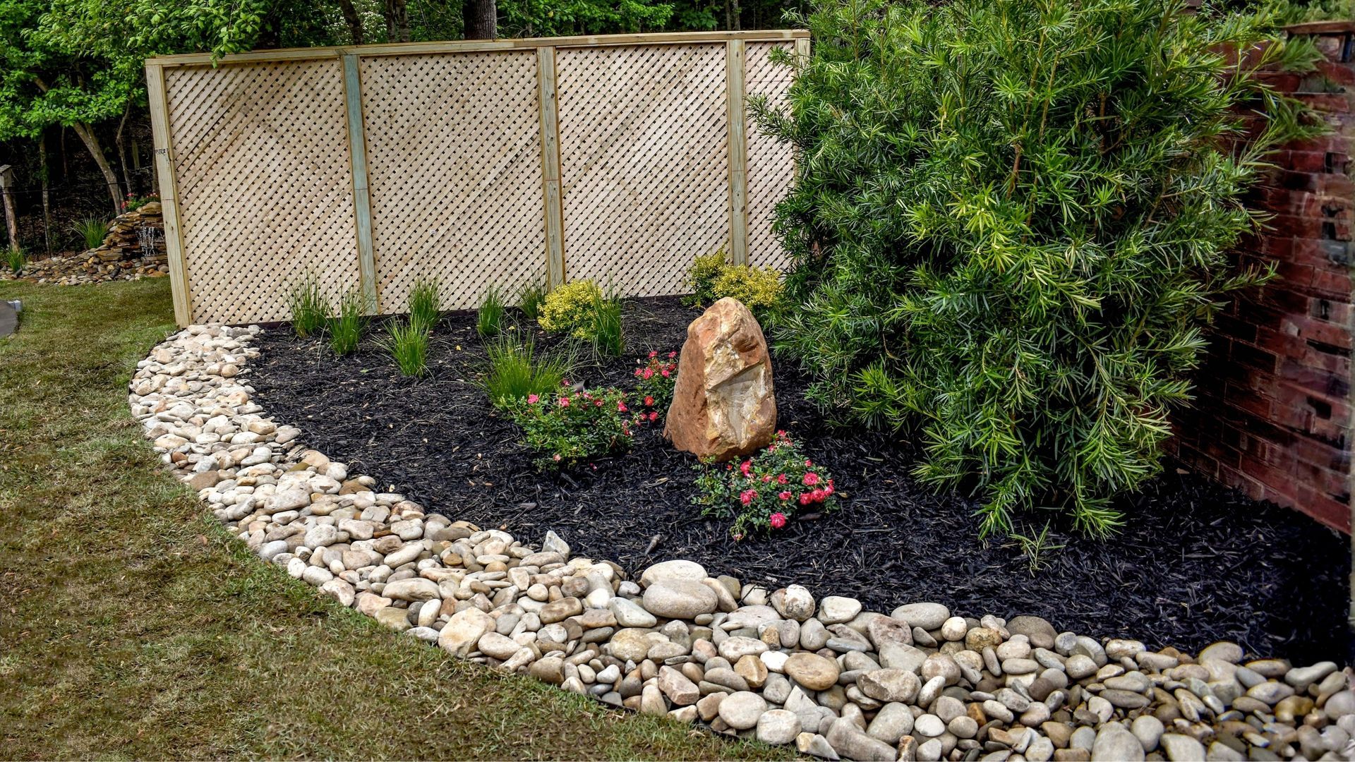 Landscaped garden bed with black mulch, white stones, shrubs, and a wooden fence.