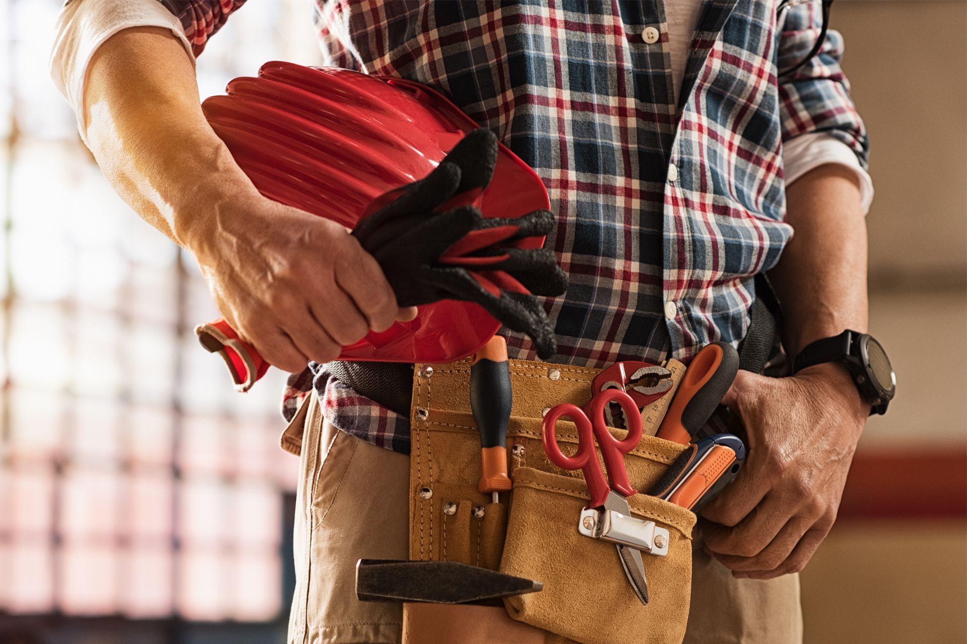 Worker holding a red hard hat beside a tool belt with hammer, pliers, and tape measure