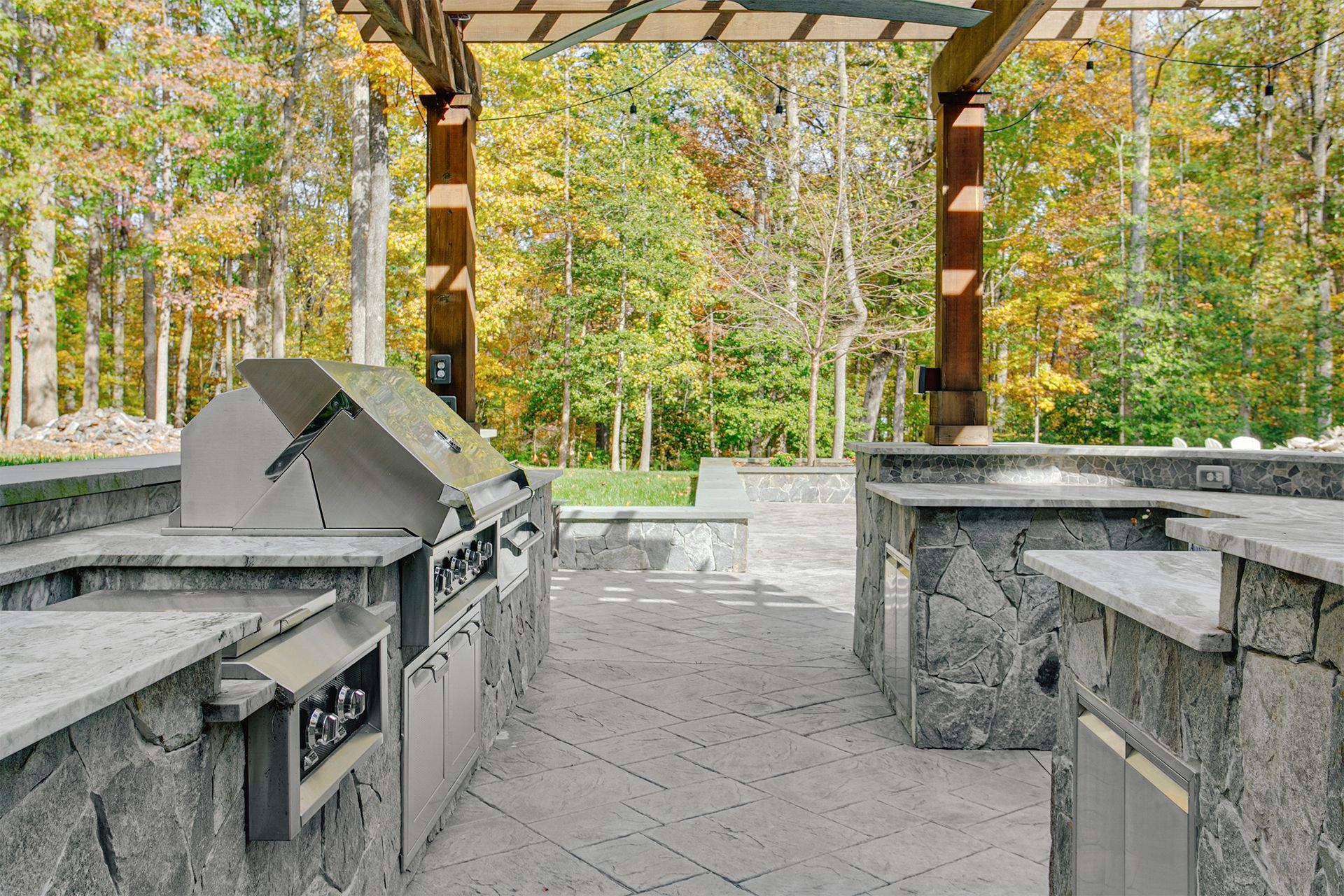Outdoor kitchen with grill, countertops, and pergola, set against a background of autumn trees.