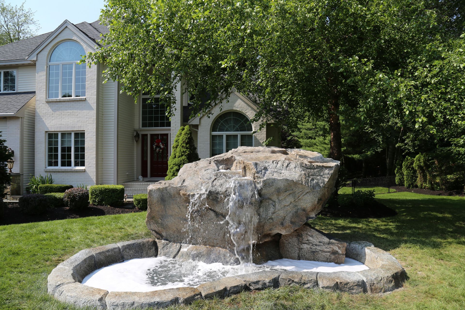 Stone fountain in front of a suburban house, with water flowing over a large boulder and circular basin.