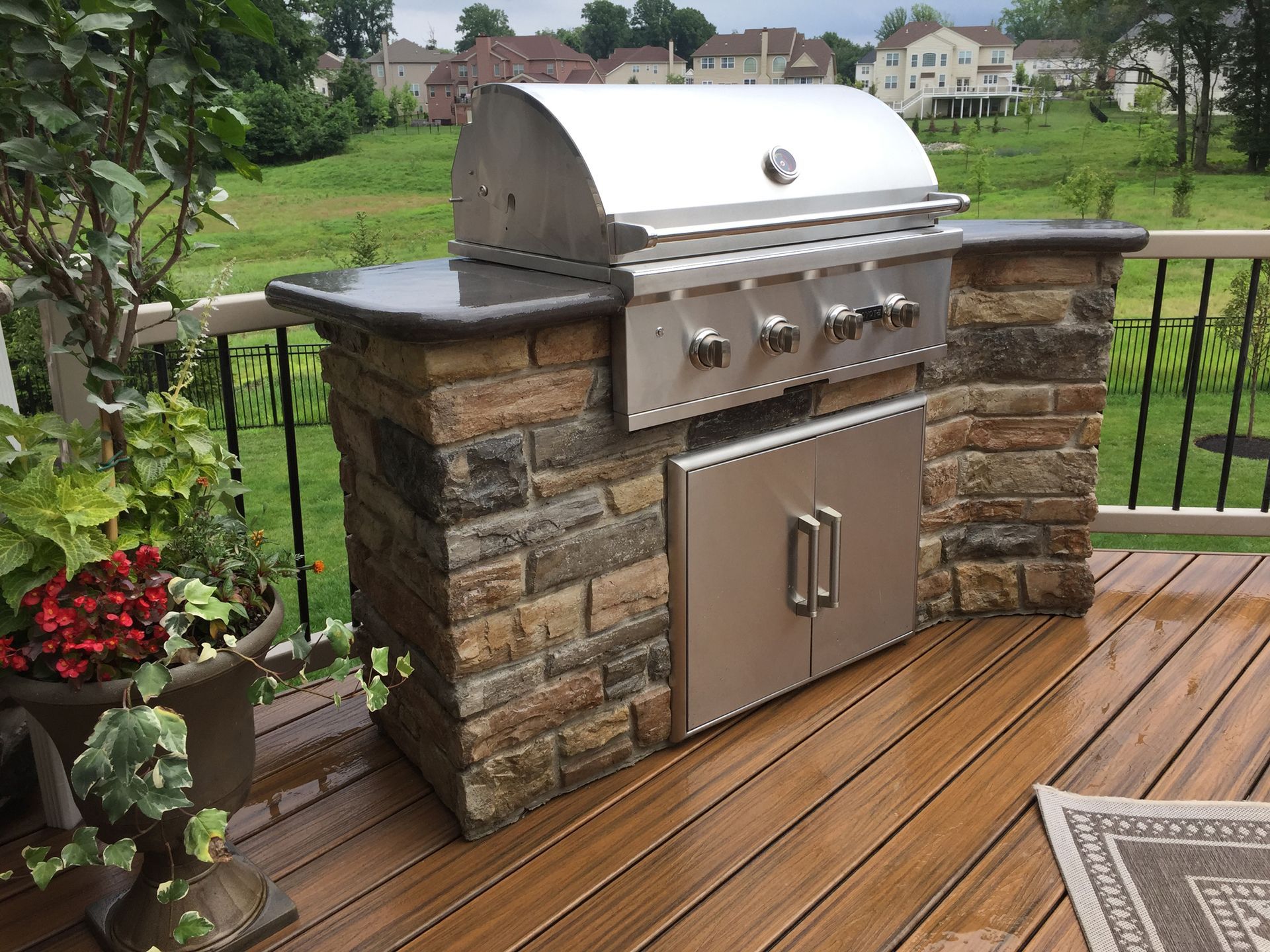 Built-in stainless steel gas grill on a wooden deck with stone sides and a grassy backyard view