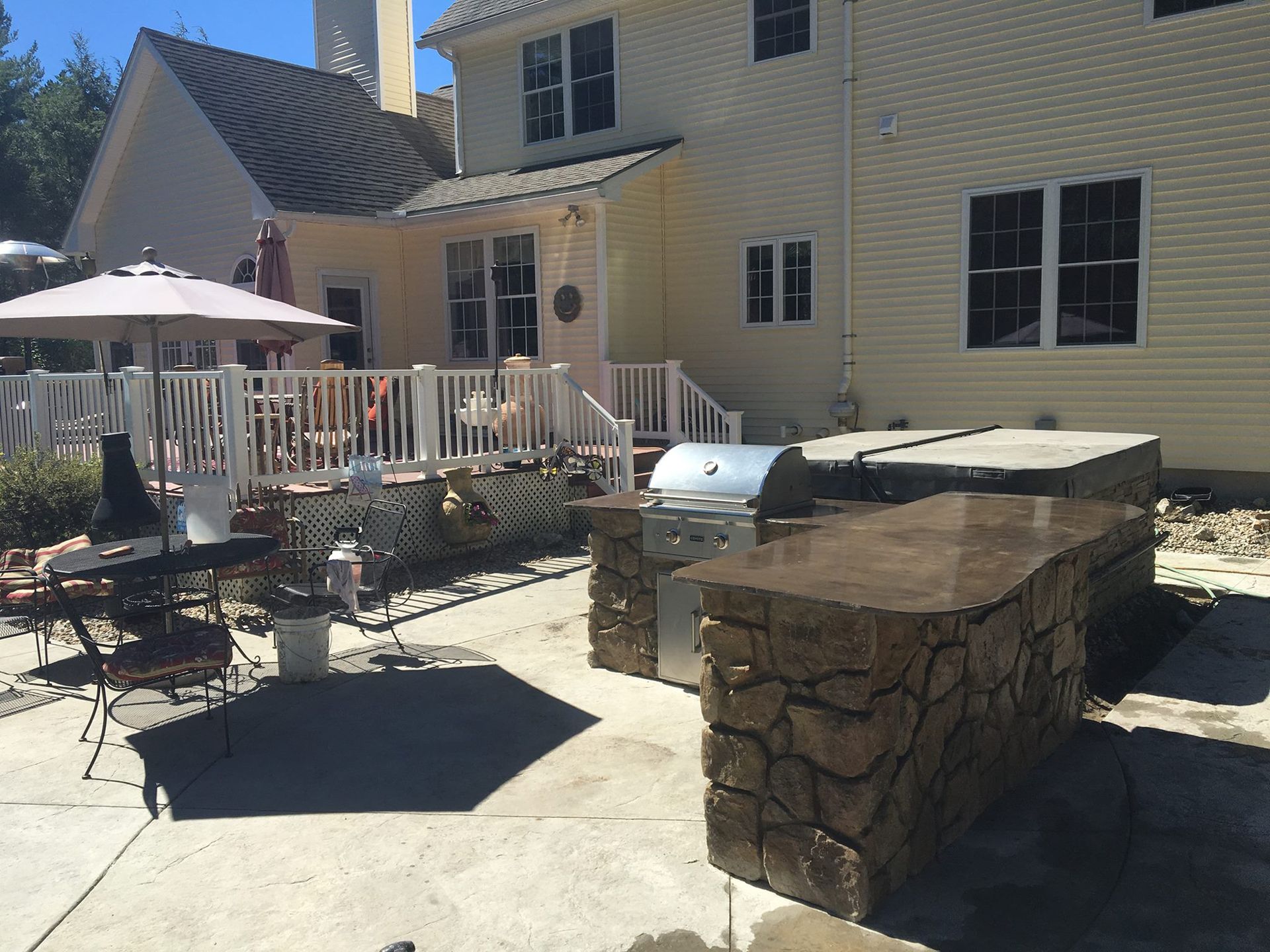Backyard patio with stone grill, table and chairs, white railing, and yellow house in sunlight