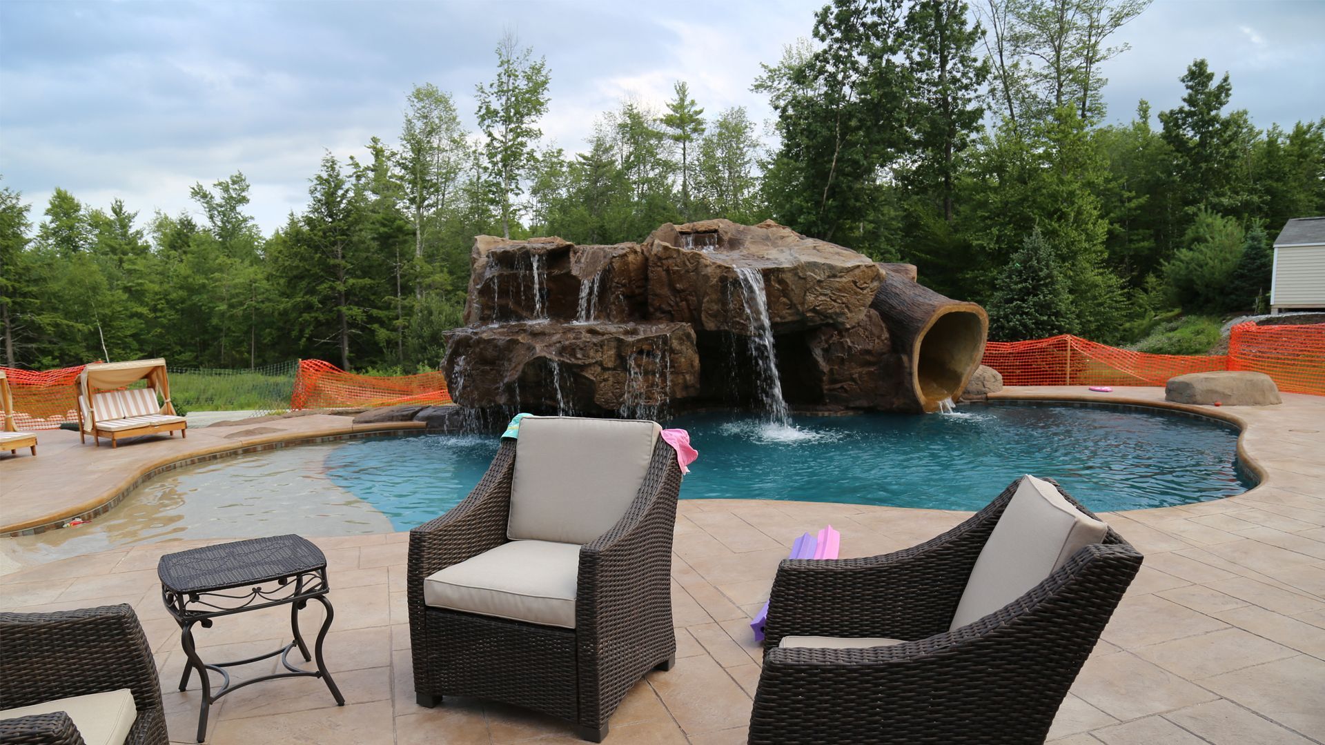 Outdoor pool with rock waterfall and wicker chairs on a wooden deck, surrounded by trees.