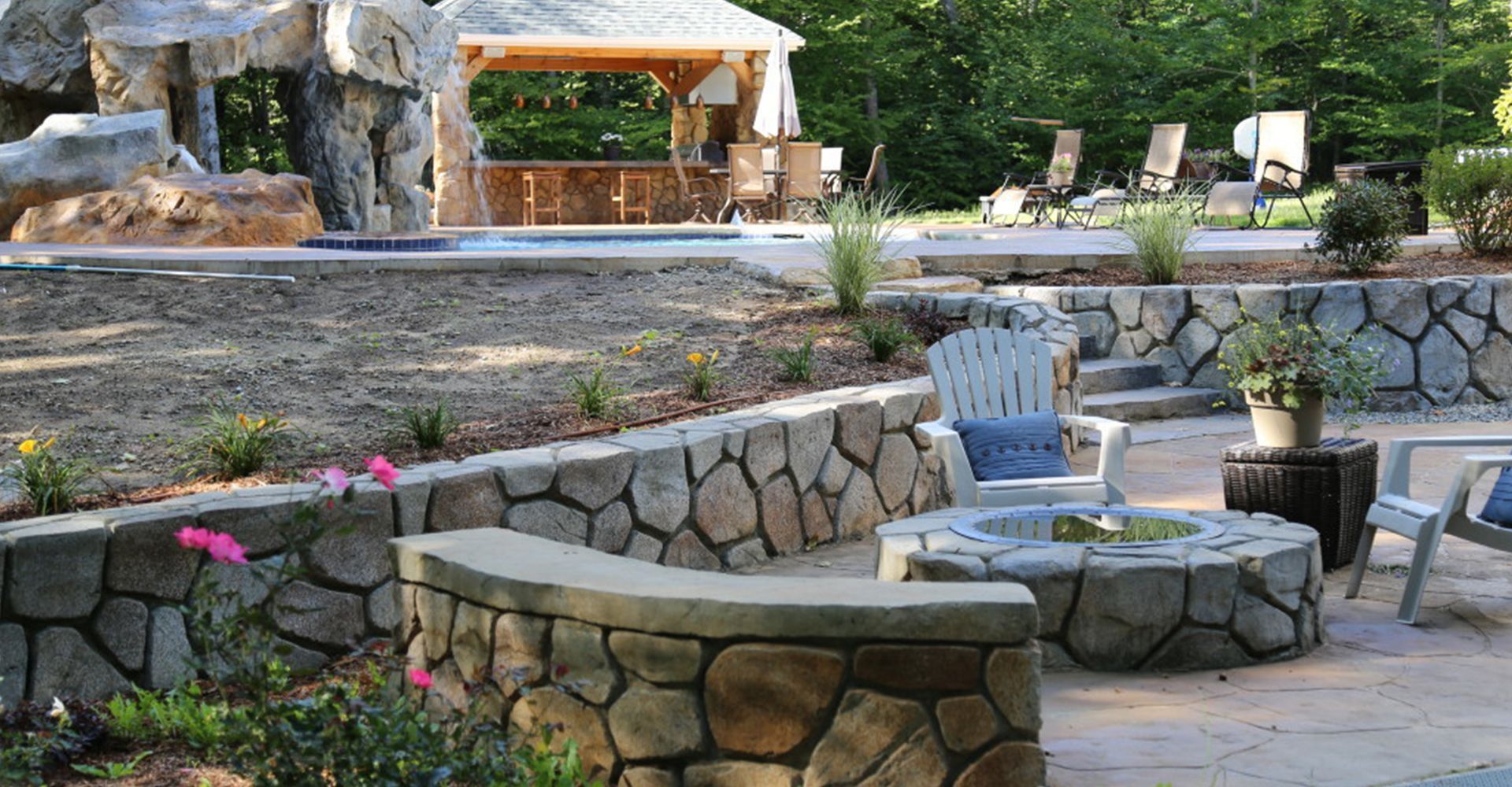 Stone patio with curved bench seating, potted flowers, and a shaded gazebo in a garden.