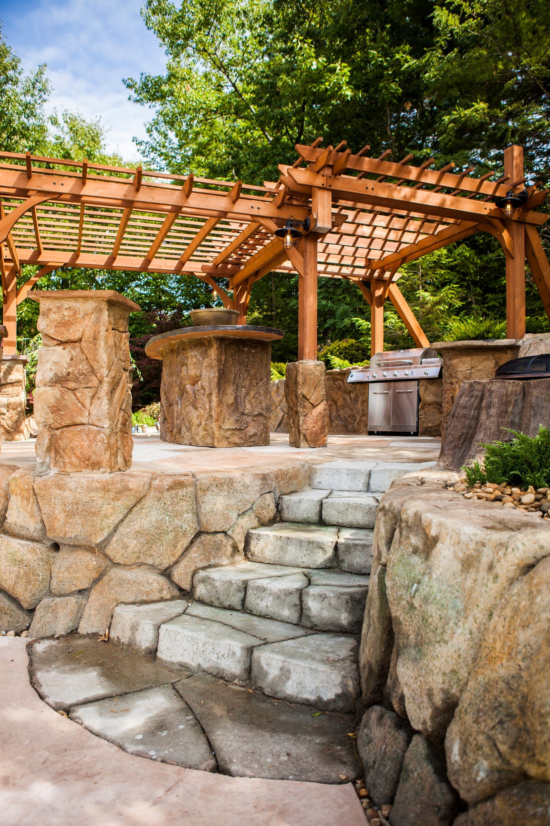 Stone steps in a landscaped garden beneath a wooden pergola, with trees and rock walls.