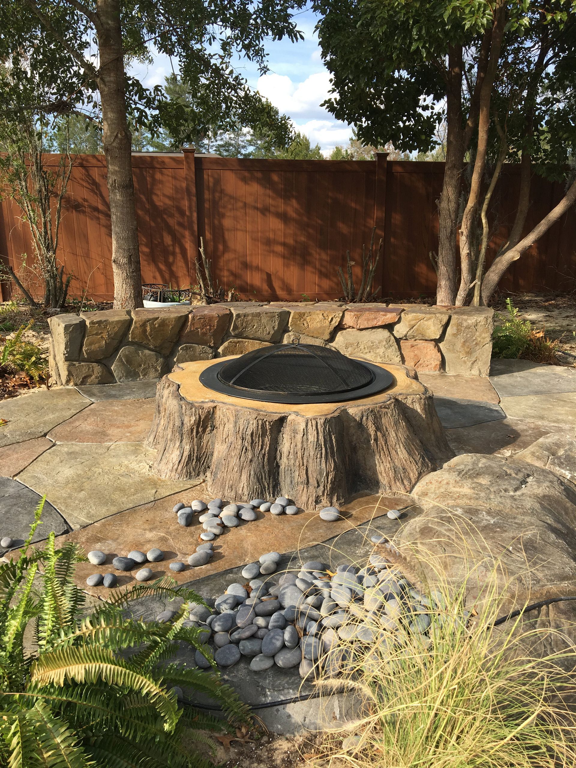 Outdoor stone fire pit with a black cover, surrounded by logs, rocks, and trees in a backyard garden