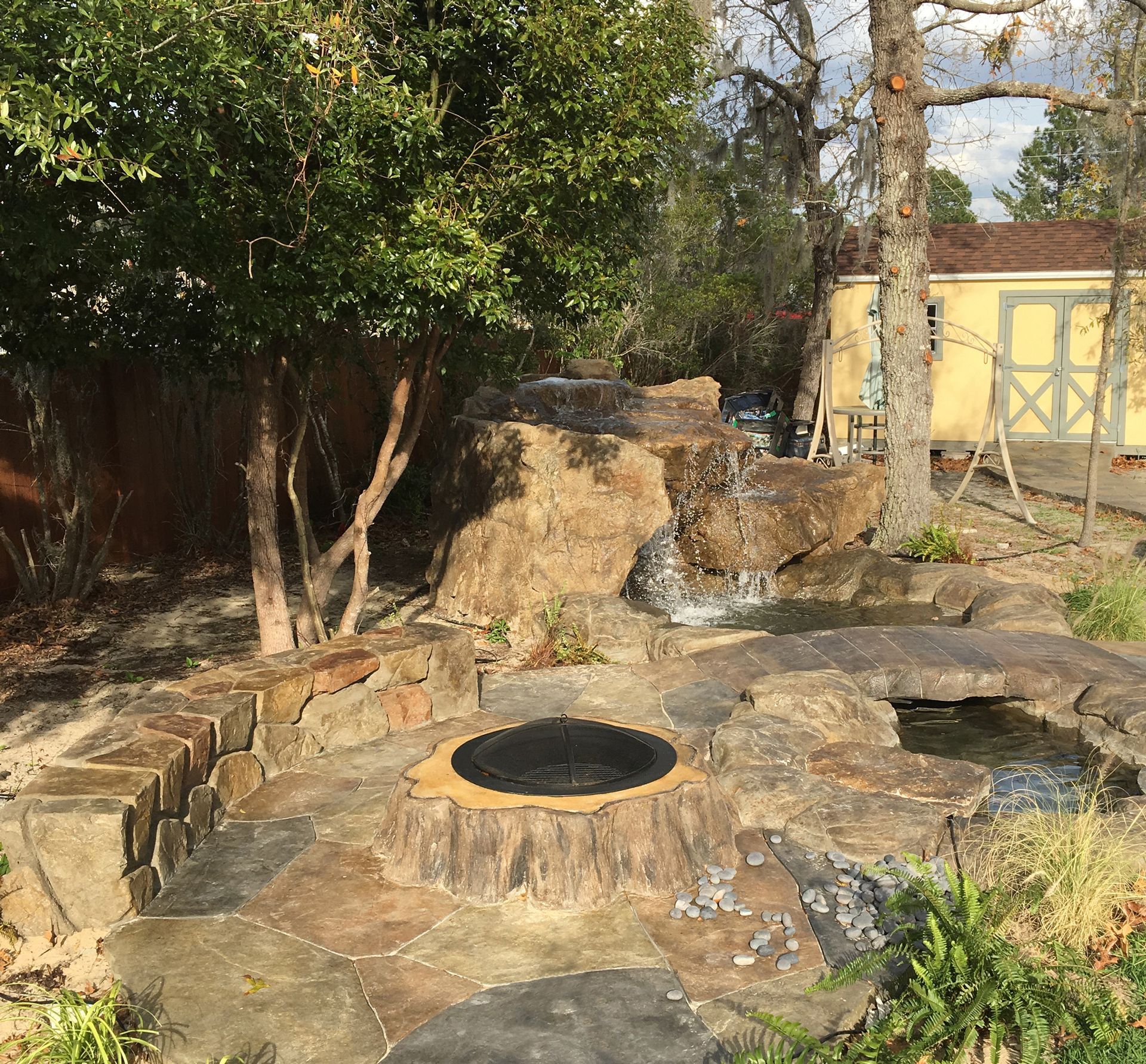Rock garden with a small waterfall, stone fire pit, and trees beside a yellow shed