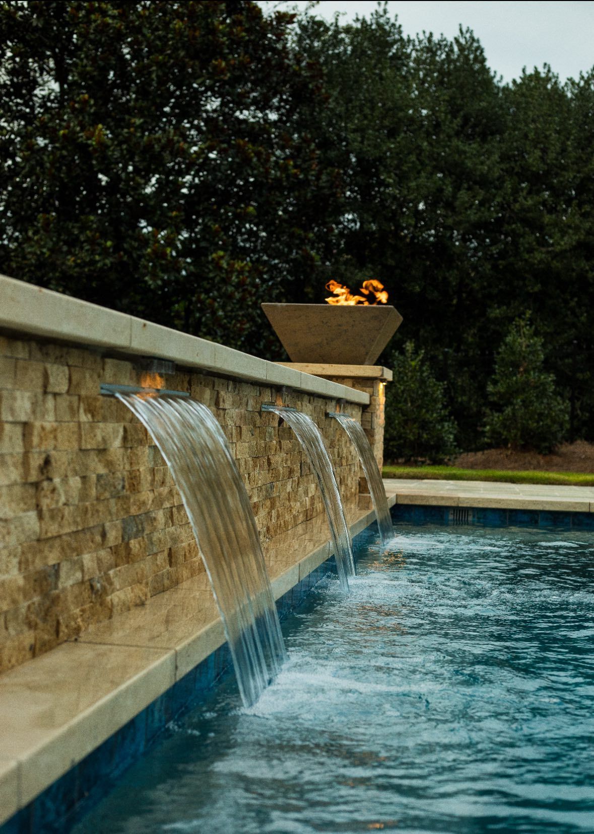 White Adirondack chairs circle a lit stone fire pit on a patio.