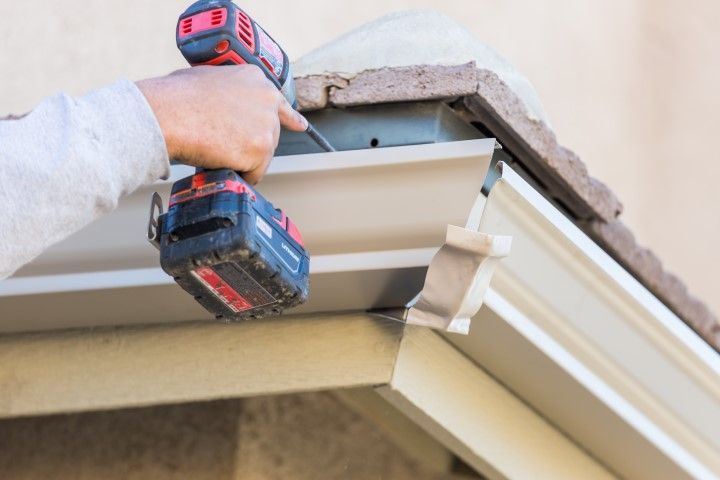 A person uses a power drill to install or repair a section of light-colored metal rain gutter under a tiled roof edge.