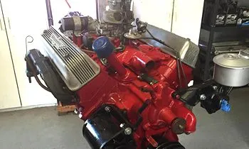 A red automotive V8 engine with finned aluminum valve covers, sitting in a garage setting.