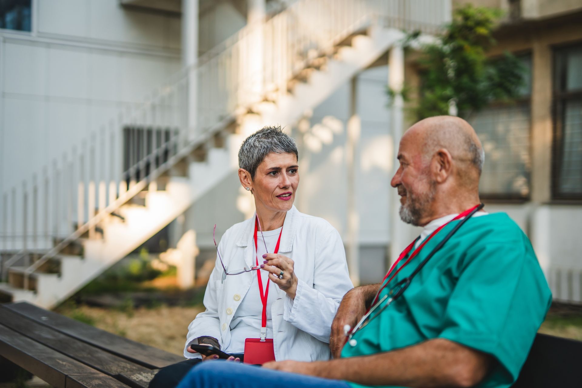 Two people chatting on a bench outdoors, one holding a cup near a staircase.