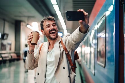 Un homme prend un selfie dans une station de métro, un café à la main, souriant à côté d'un train.