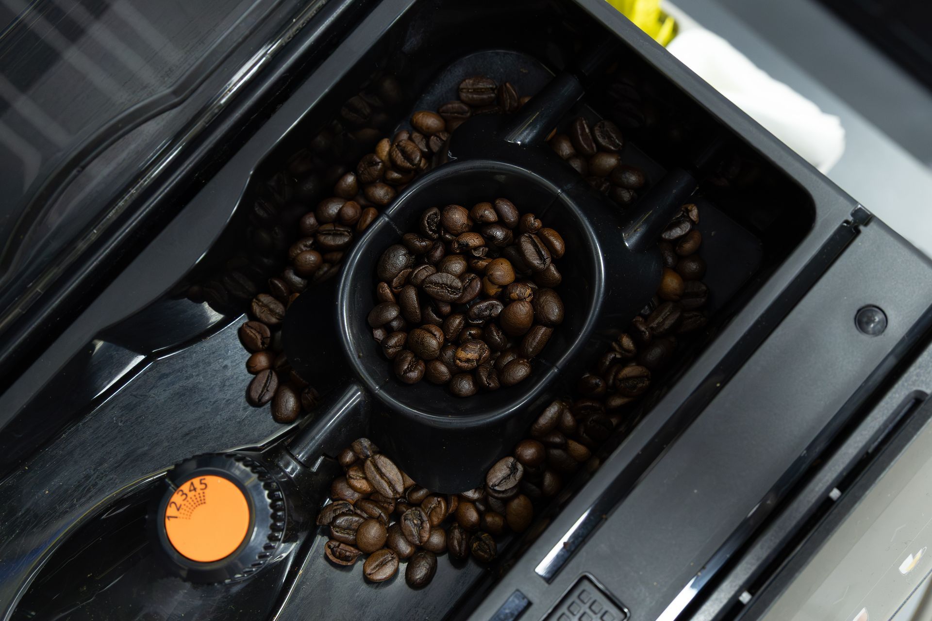 Coffee beans in a machine’s hopper, viewed from above, with a black and orange control knob.