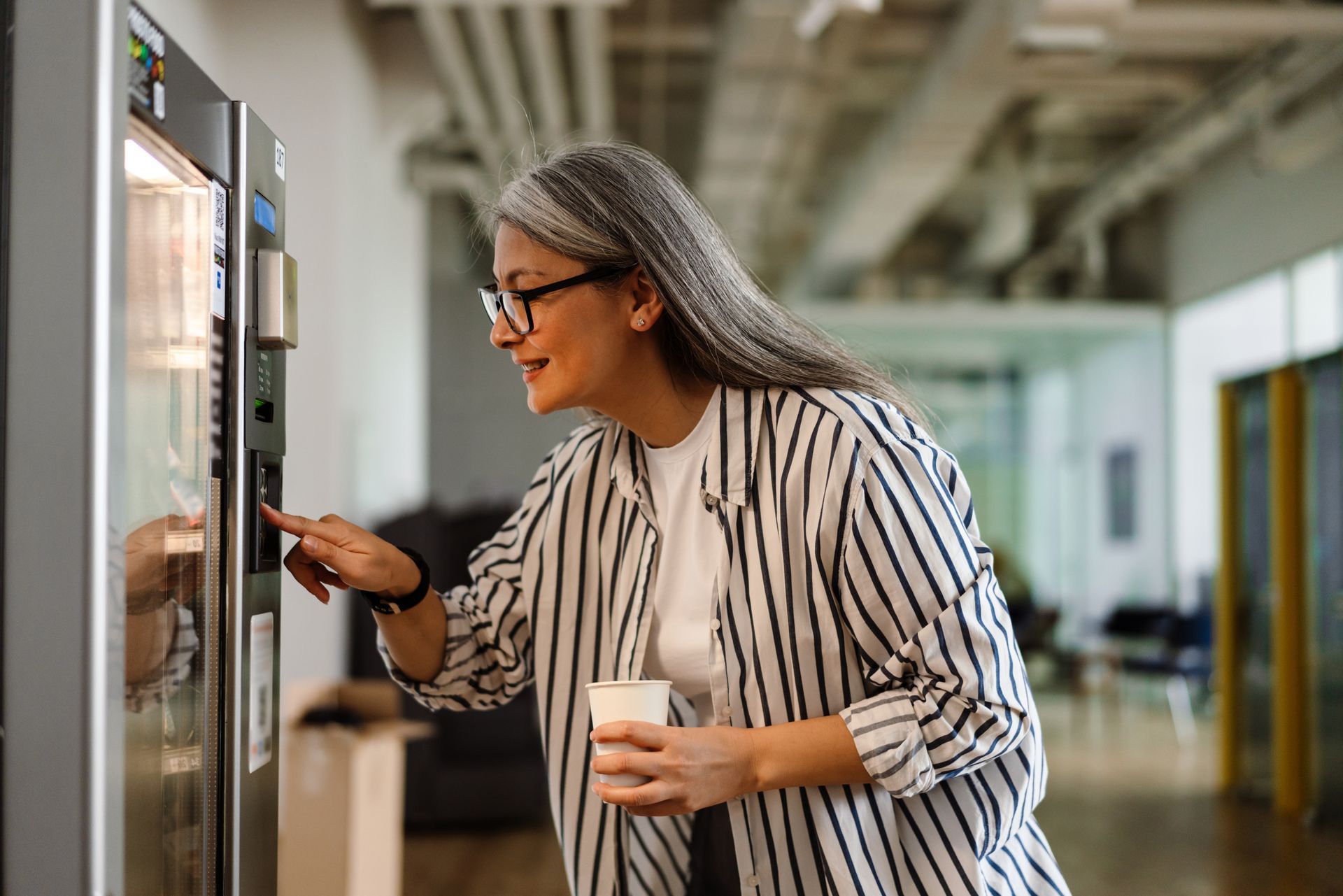 Une femme appuie sur un bouton d'un distributeur automatique tout en tenant une tasse, dans un environnement de bureau.