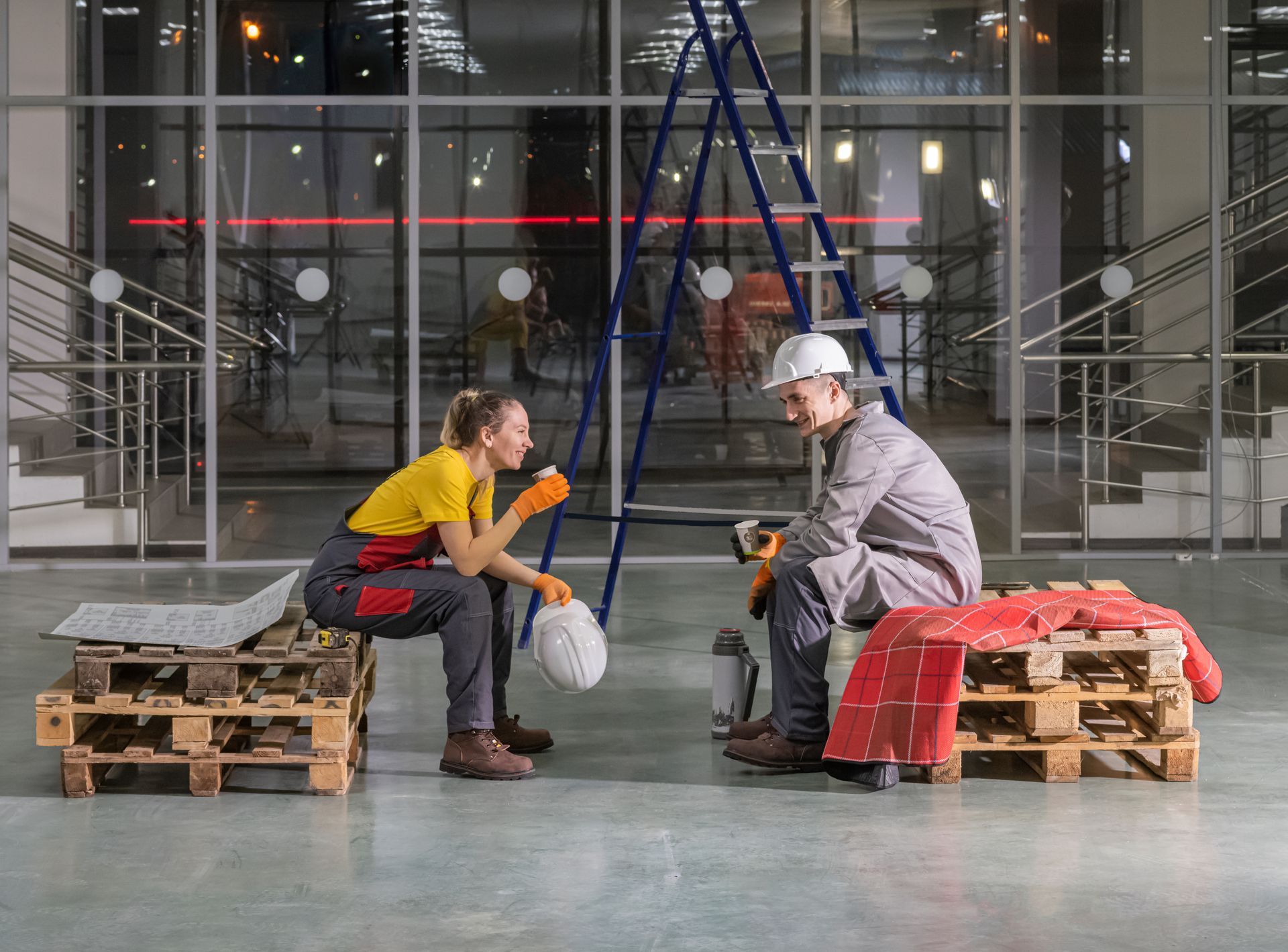 Two workers in hard hats sit on pallets in a construction site, facing each other with tools.
