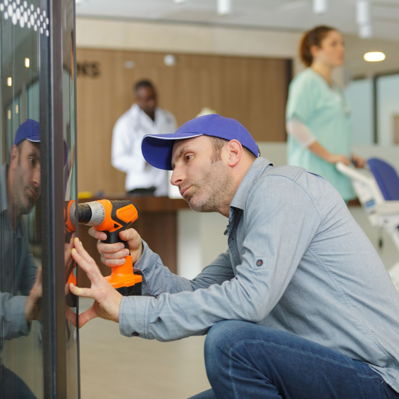 Un technicien utilise une perceuse orange pour réparer une vitre dans un hall d'entrée.