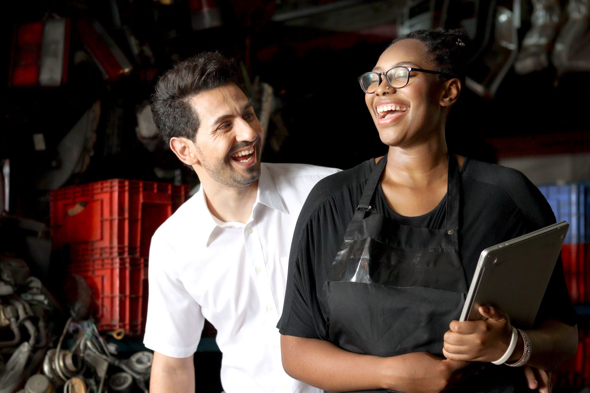 Two smiling coworkers in an industrial setting, one holding a tablet and wearing an apron.