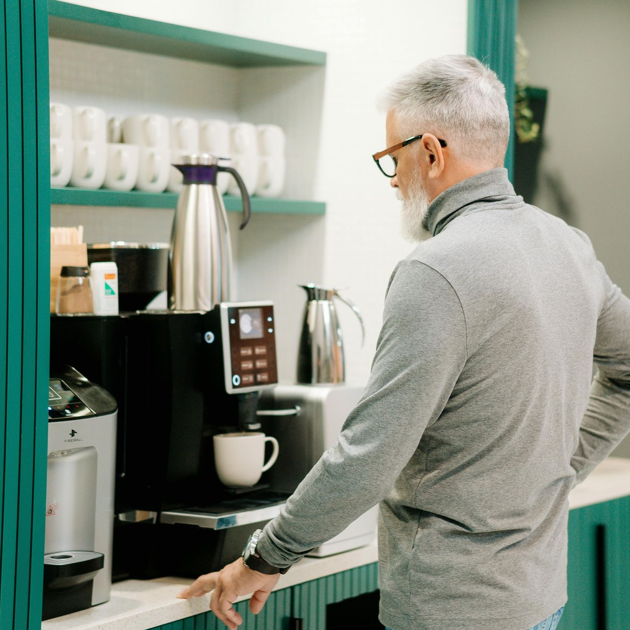 Un homme en pull gris utilise une machine à café à un comptoir de cuisine turquoise.
