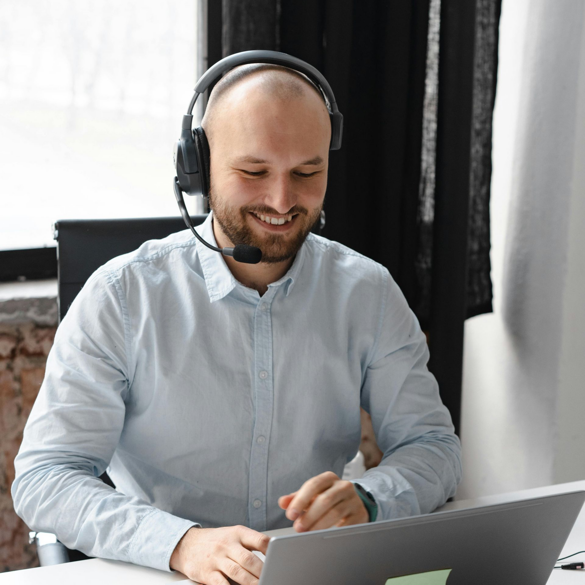 Un homme souriant, portant un casque audio et parlant sur un ordinateur portable, dans un bureau lumineux