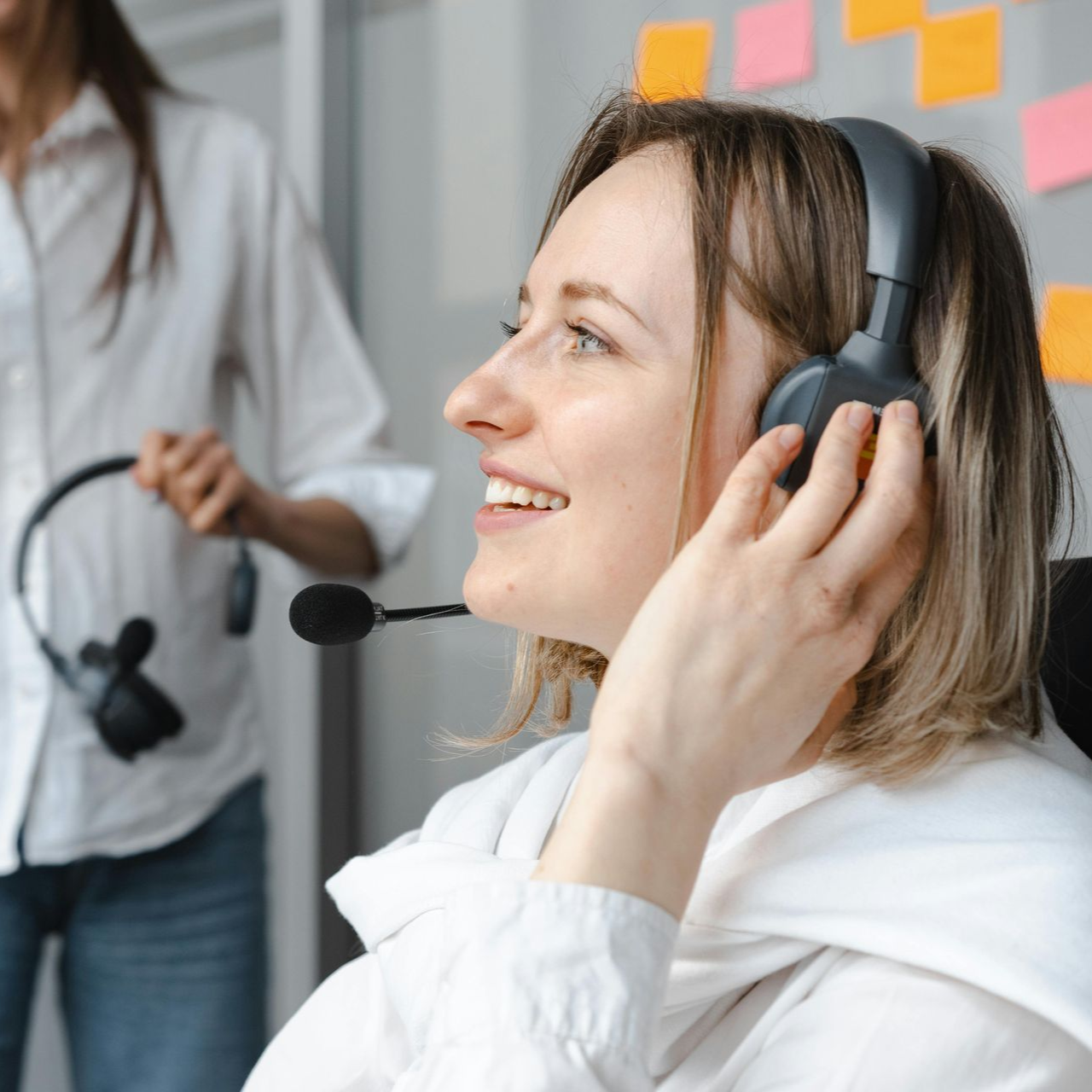 Une femme portant un casque audio sourit dans un bureau, une autre personne étant floue en arrière-plan.