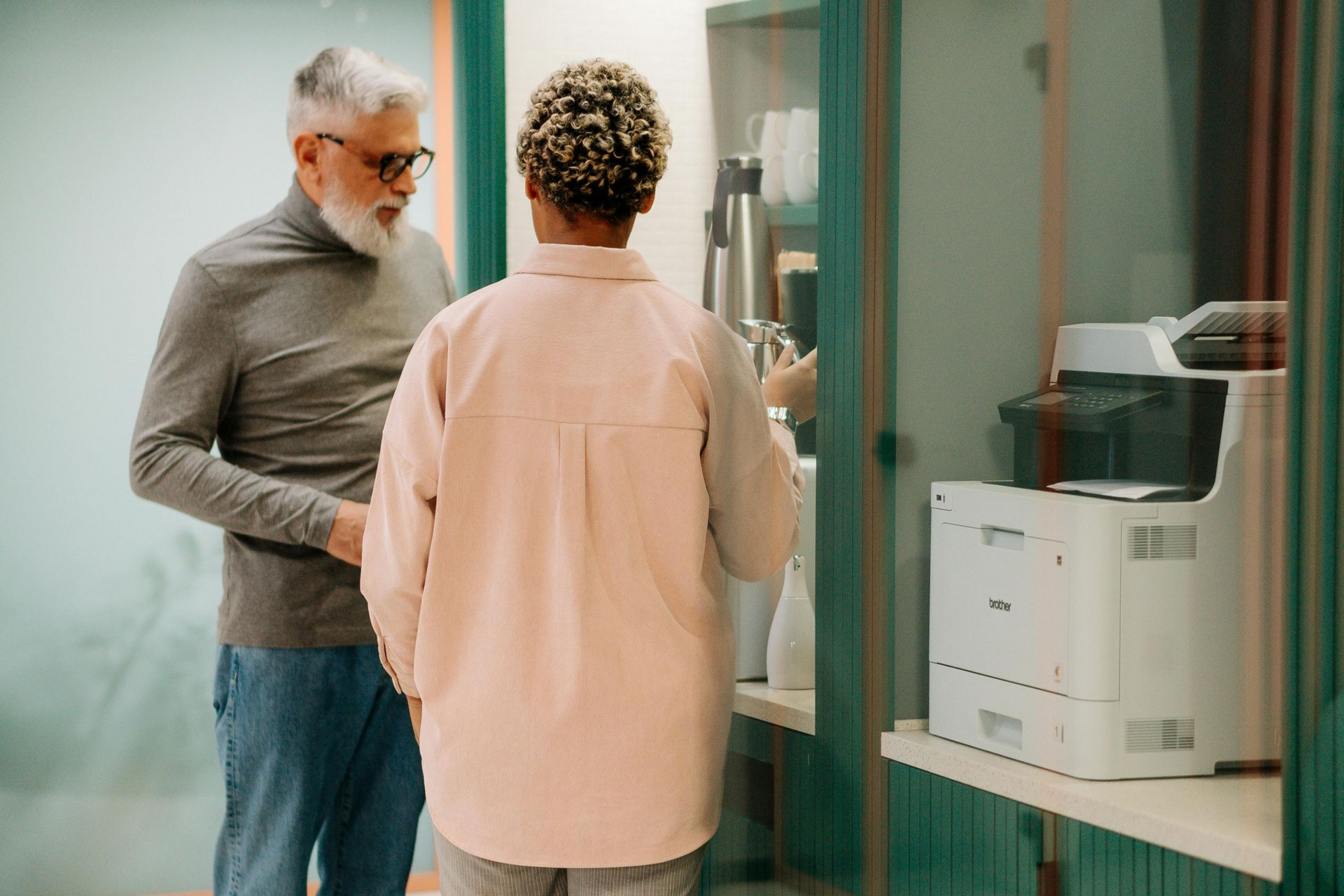 Two people talking in a hallway beside a glass office door and copier.