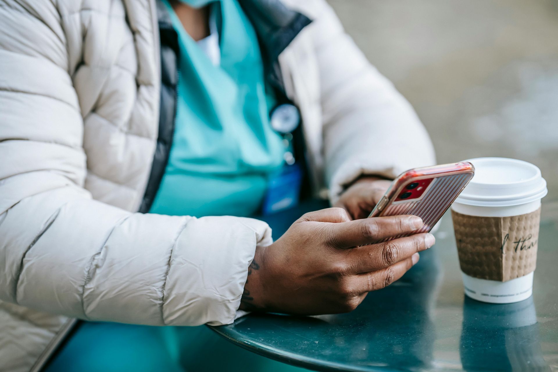 Une personne en veste blanche utilise un smartphone à côté d'une tasse de café à emporter, à une table.