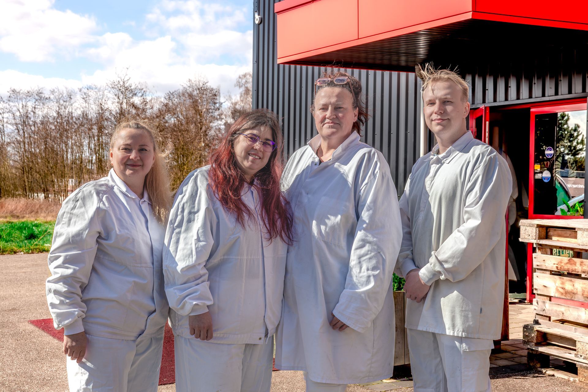 Quatre personnes en vêtements de travail légers posent devant un bâtiment rouge, au soleil.