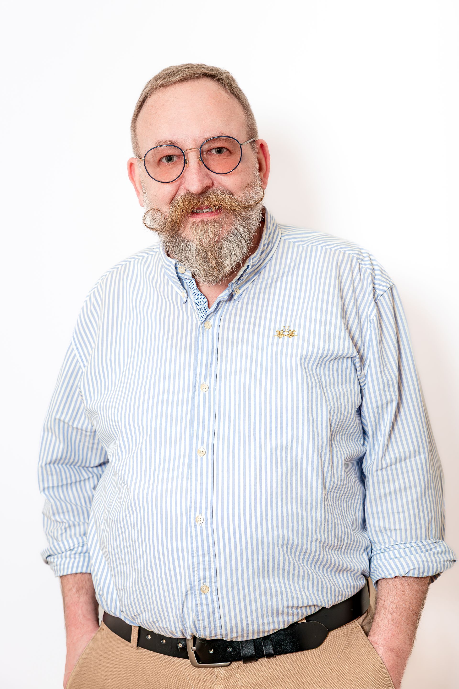 Homme souriant portant des lunettes et une barbe, vêtu d'une chemise à rayures bleu clair sur fond blanc