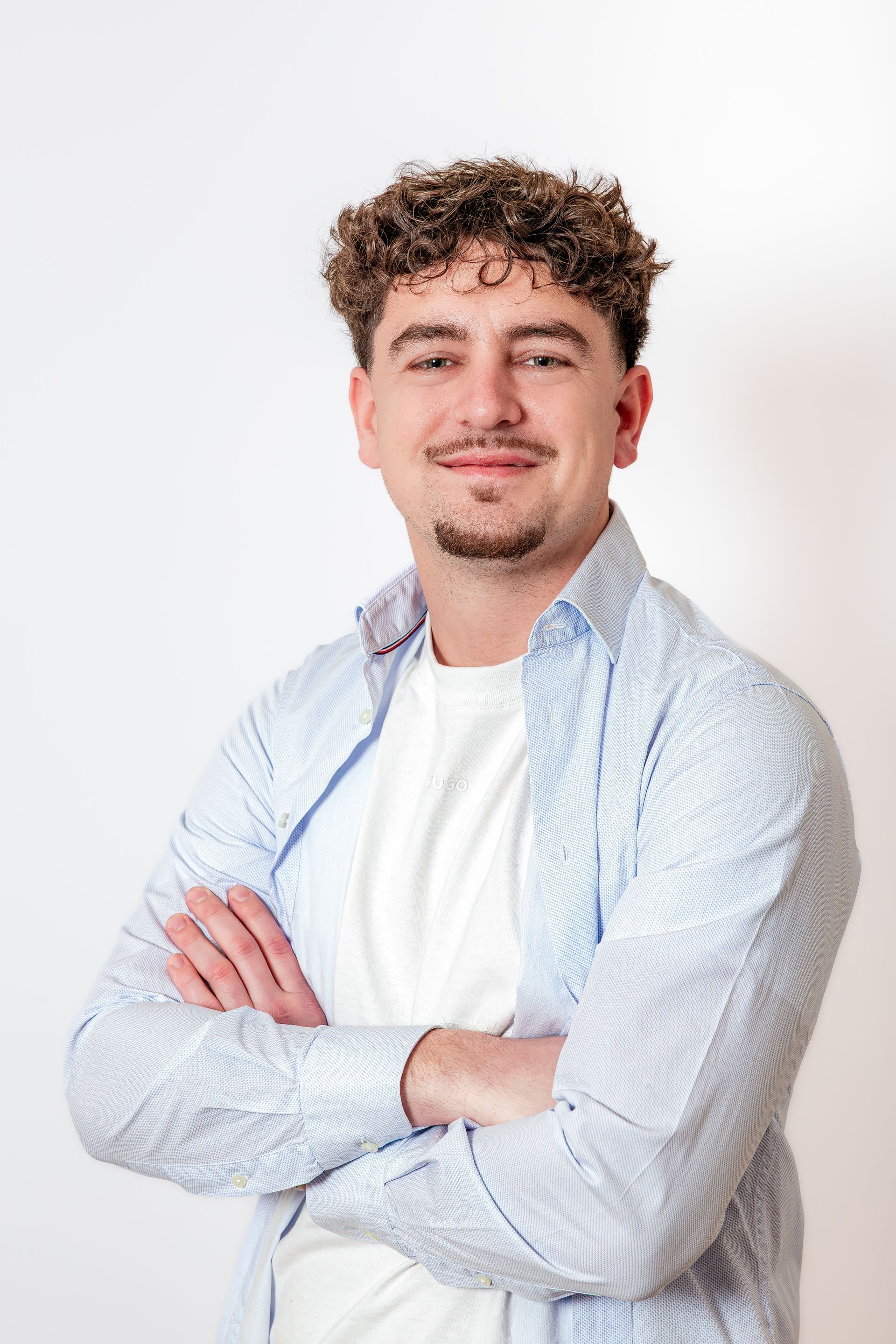Homme debout, les bras croisés, souriant, vêtu d'une chemise bleu clair et d'un t-shirt blanc, sur fond uni.