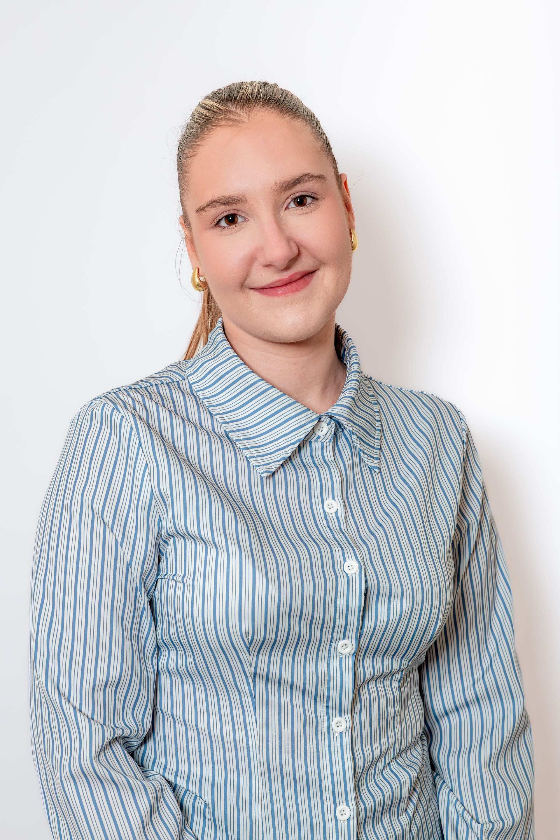 Personne portant une chemise bleue à rayures boutonnée, souriant sur un fond blanc uni.