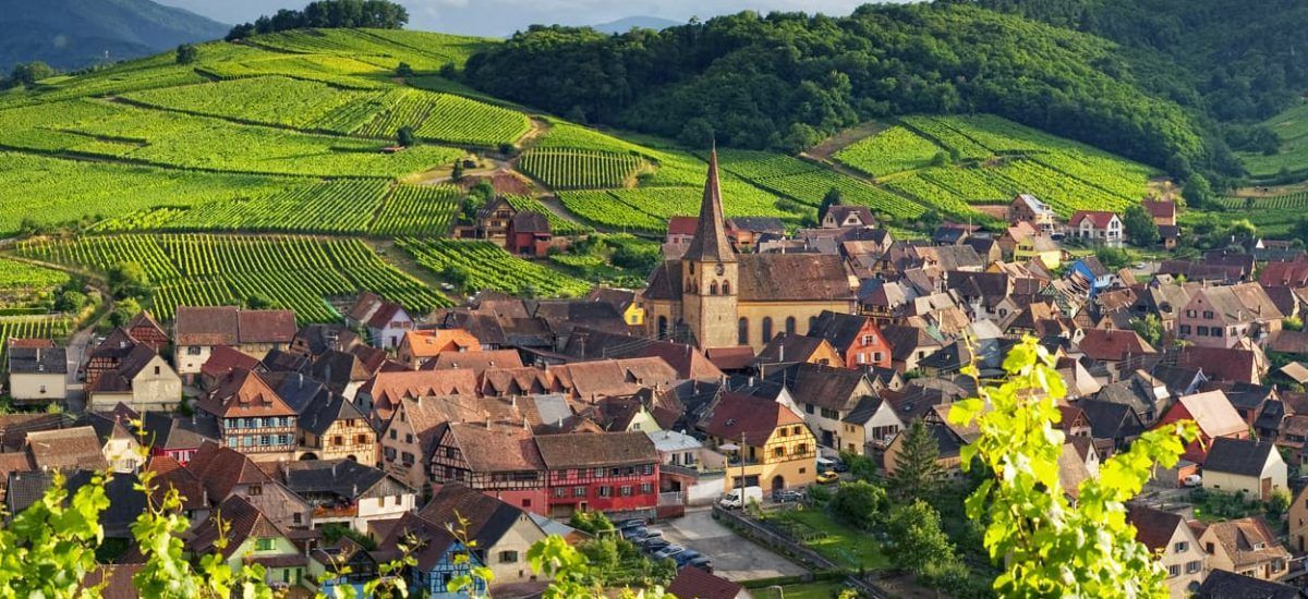 Village with a church tower beside green vineyard-covered hills under bright sunlight