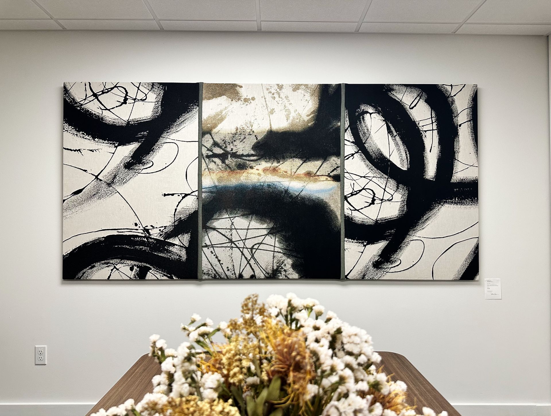 A large black and white painting is hanging on a wall above a table with flowers.