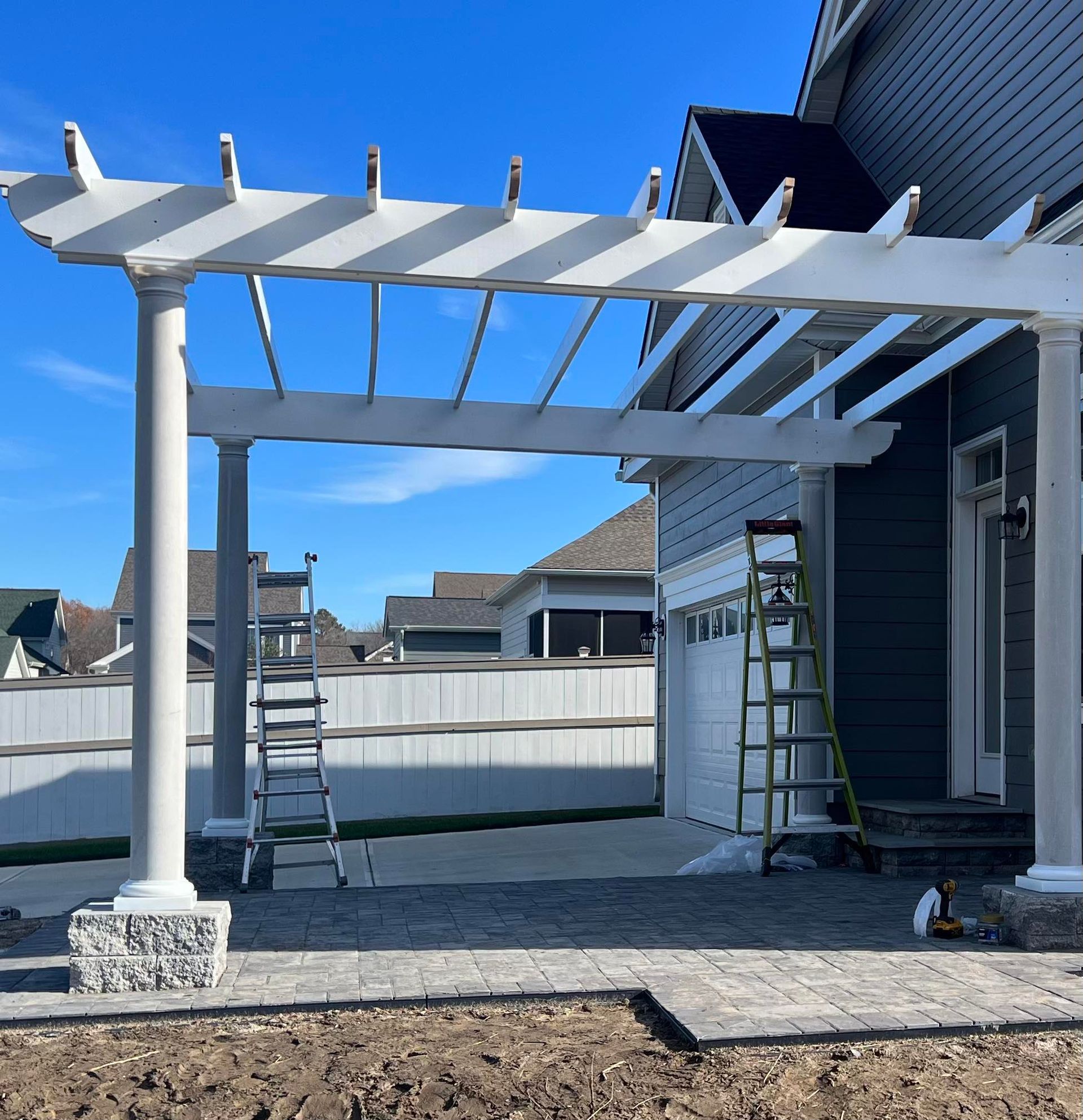 Backyard patio pergola being built beside a house, with ladders and scaffolding under a blue sky