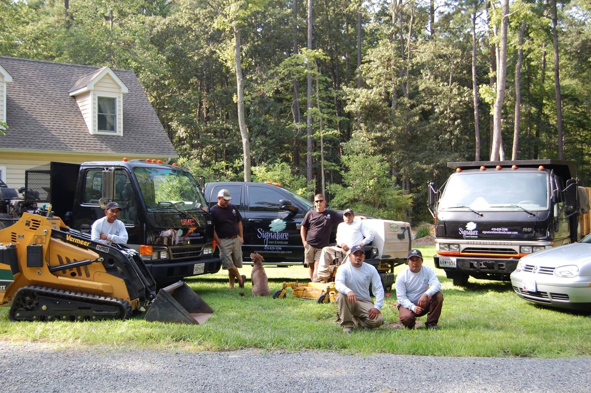 People and equipment gathered on a grassy driveway beside a house and trucks in a wooded yard