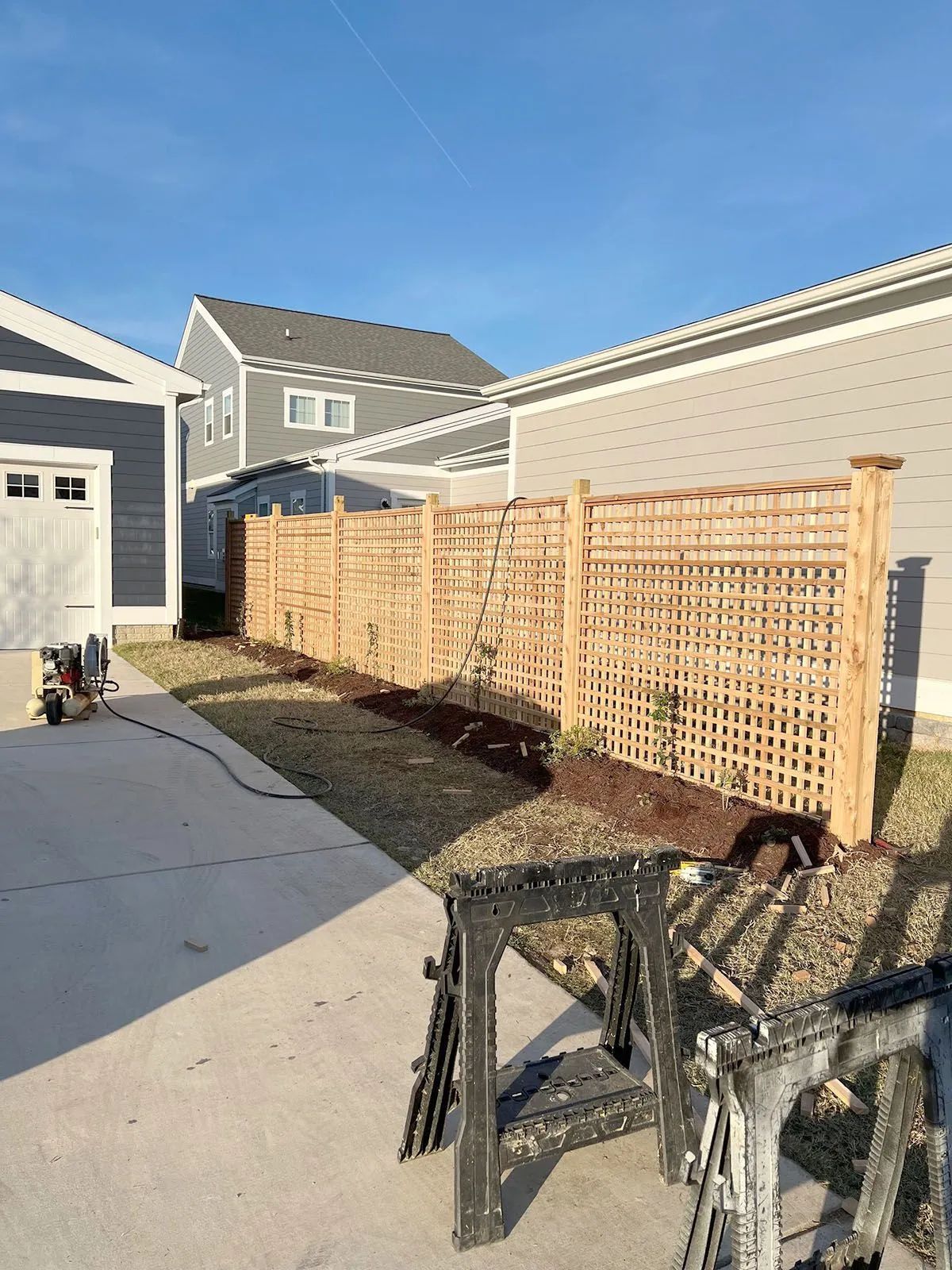 Residential backyard with a wood fence, concrete driveway, and sawhorse in the foreground under a blue sky