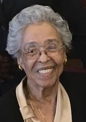 Smiling elderly woman with white hair and glasses, wearing a beige blouse against a dark background