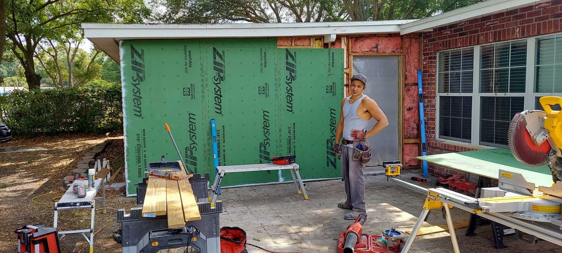 Person standing beside a partially covered green wall during outdoor home renovation, with tools and lumber around.