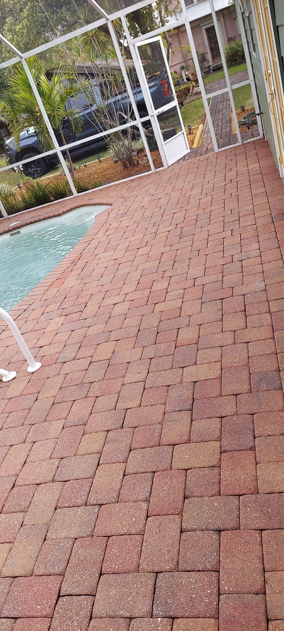 Brick patio beside a pool, with white fencing and greenery in the background.