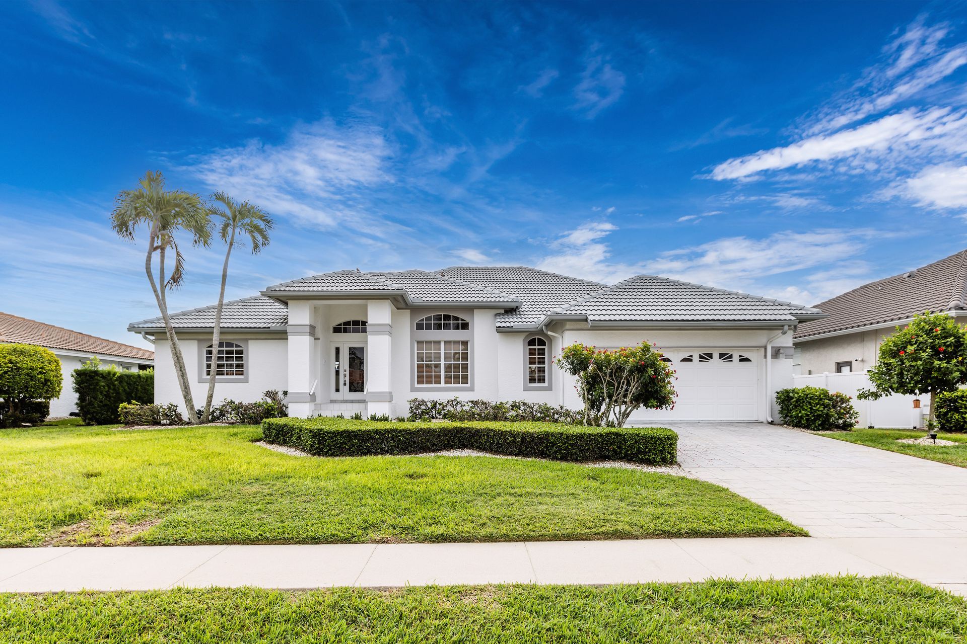 Single-story white suburban house with a two-car garage, green lawn, and palm trees under a blue sky Single-story white suburban house with a two-car garage, green lawn, and palm trees under a blue sky