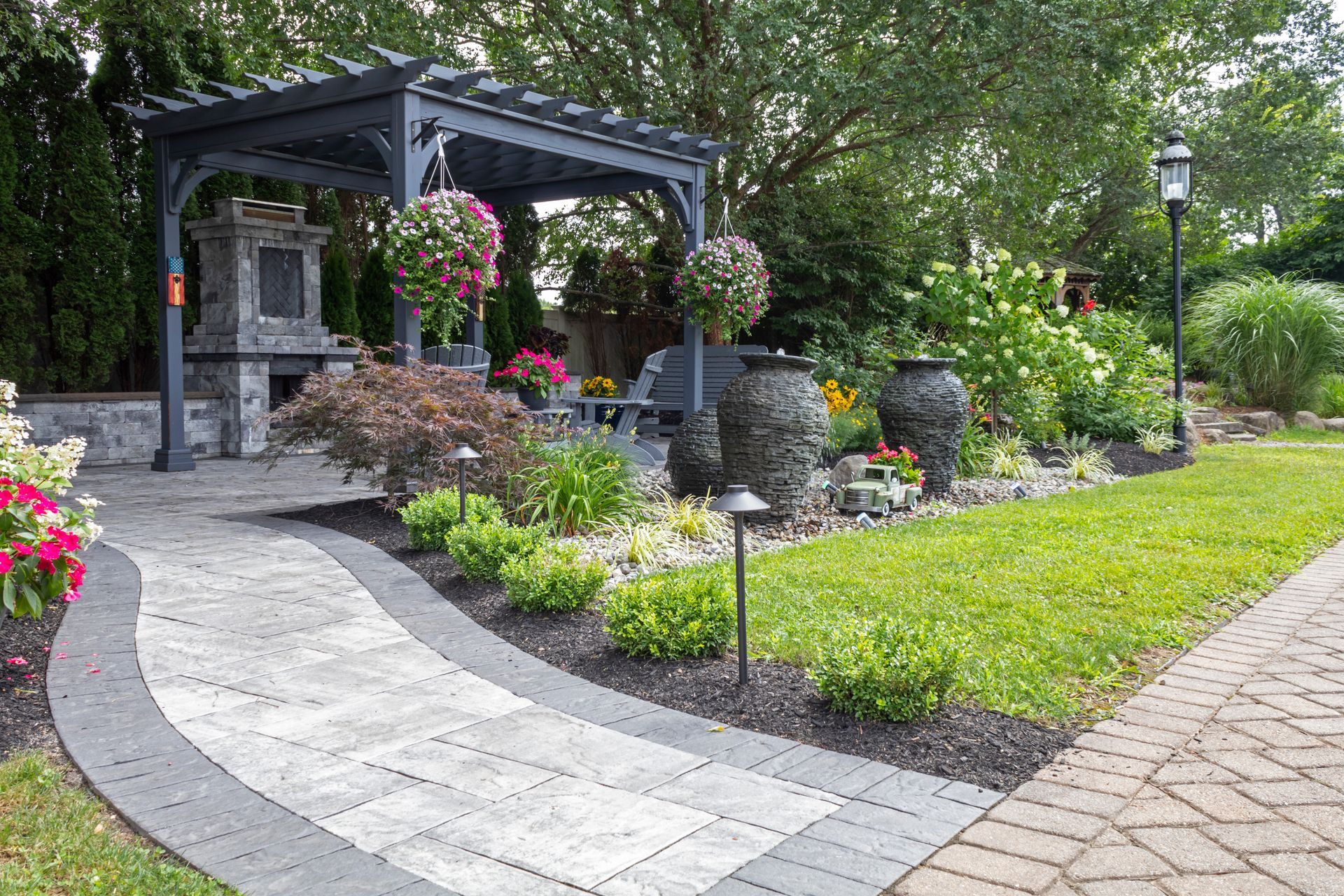 Stone patio garden with a black pergola, hanging flowers, and colorful landscaping along a curved walkway Stone patio garden with a black pergola, hanging flowers, and colorful landscaping along a curved walkway