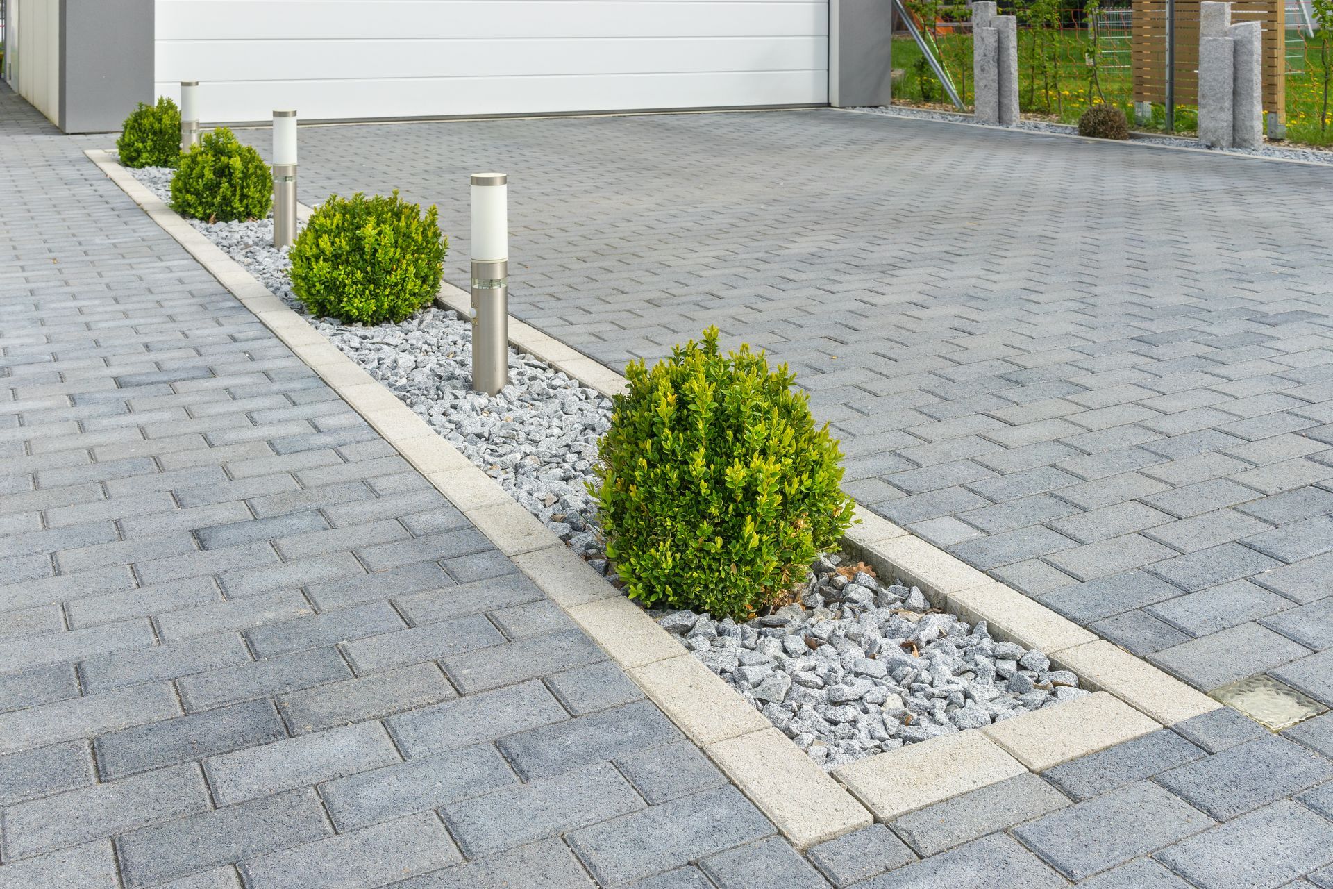 Paved driveway with gray pavers, white gravel strip, small shrubs, and a garage door in the background Paved driveway with gray pavers, white gravel strip, small shrubs, and a garage door in the background