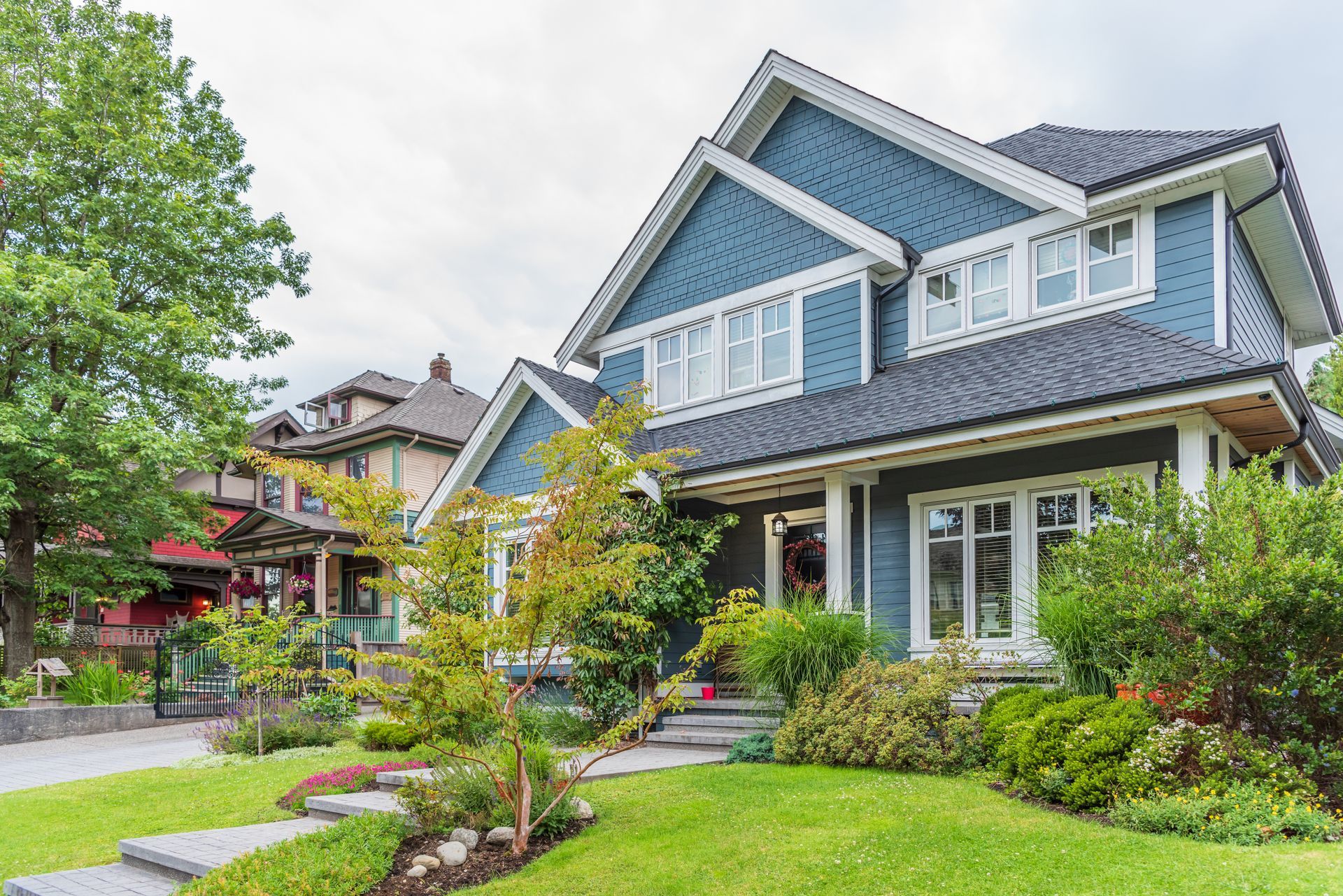 Blue-gray suburban house with white trim, landscaped front yard, and tree-lined street. Blue-gray suburban house with white trim, landscaped front yard, and tree-lined street.