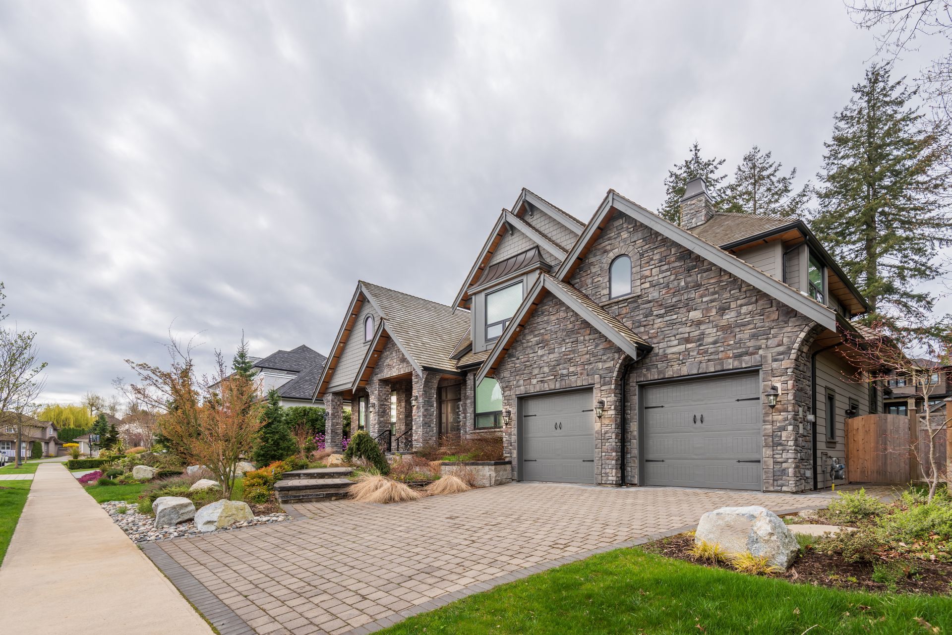 Modern stone-and-wood suburban house with a garage, front walkway, and landscaped yard under cloudy skies Modern stone-and-wood suburban house with a garage, front walkway, and landscaped yard under cloudy skies