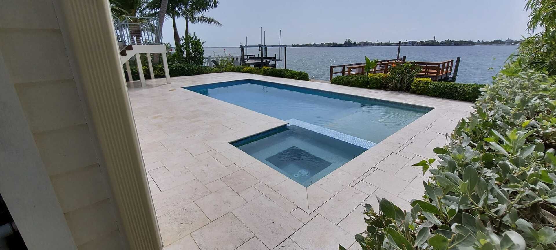 Patio with a rectangular pool and spa overlooking a waterfront, bordered by greenery and white tile