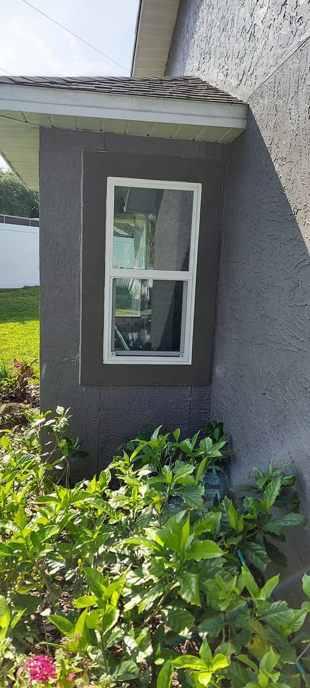 Small white-trimmed window on a gray house exterior, partly framed by green shrubs and roof eave