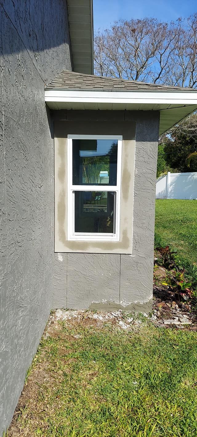 Small white-framed window on a gray stucco wall beside a lawn and trees