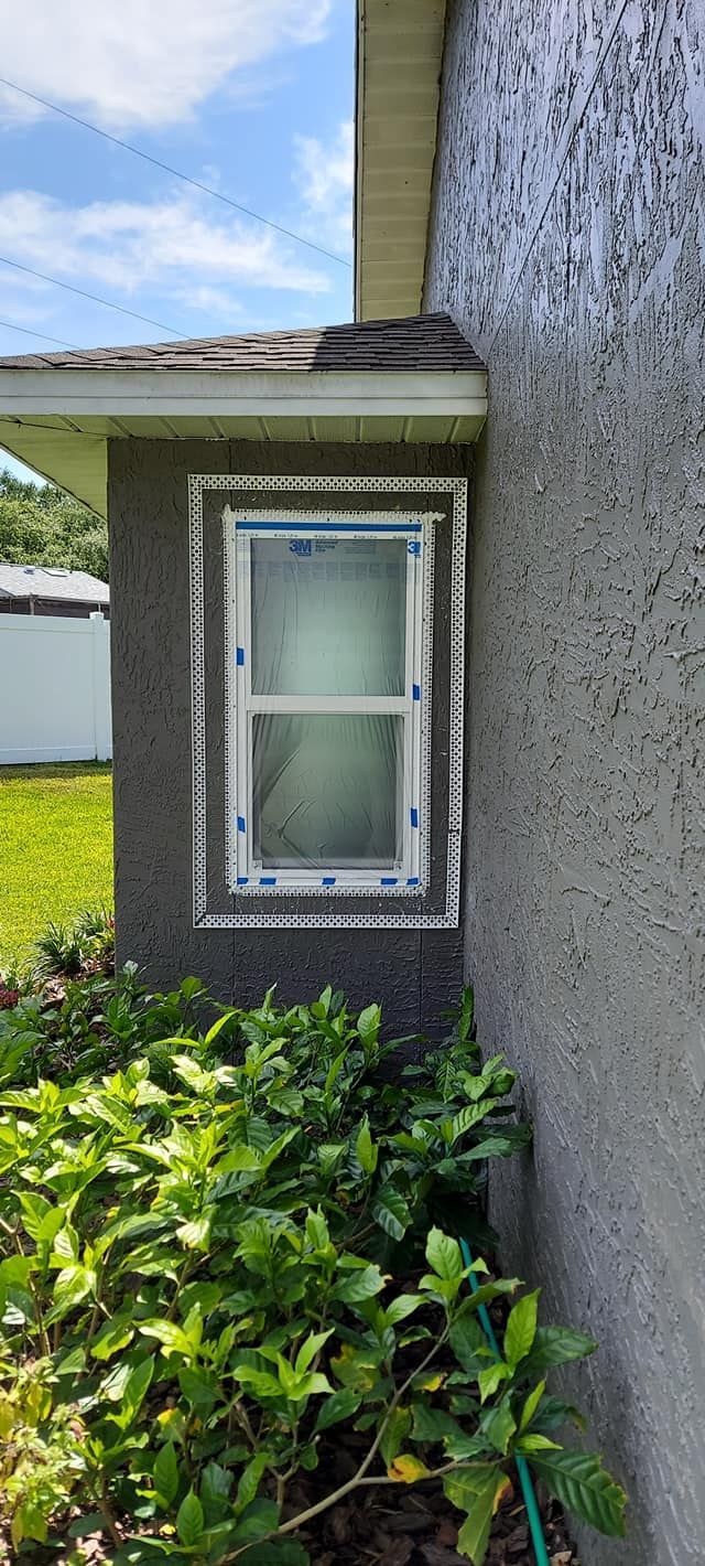 Side view of a house exterior with a window, stucco wall, and green shrubs below.