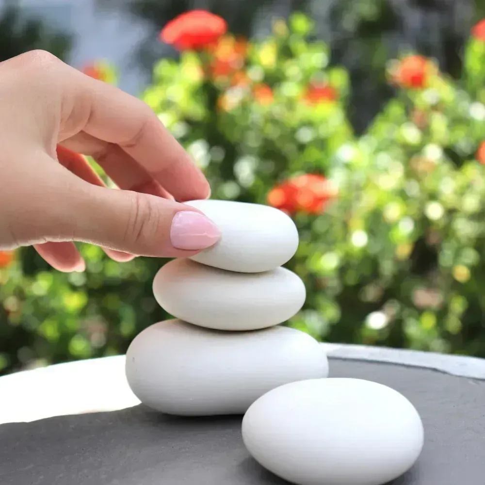 A hand stacks smooth white stones on a dark surface, with blurred green foliage and red flowers in the background.